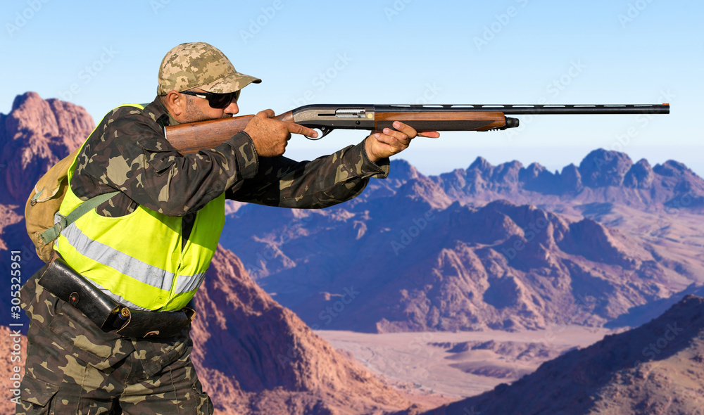 Fototapeta premium A hunter in camouflage and a gun in his hands in the early morning on top of a mountain stands looking for a target. Beautiful mountain landscape, the opening of the hunt.