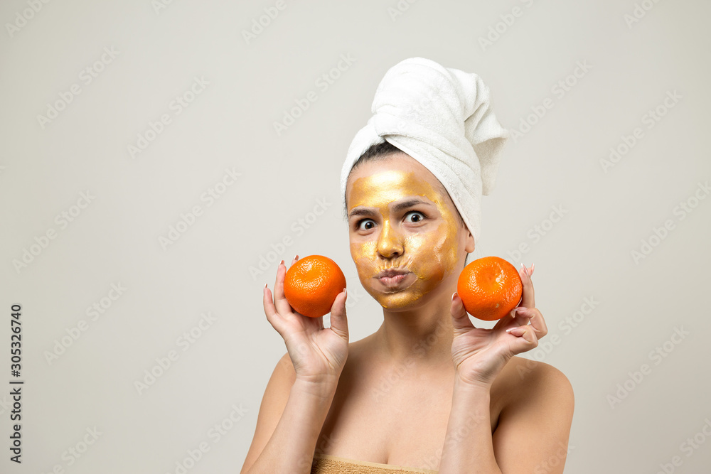 Beauty portrait of woman in white towel on head with gold nourishing mask on face. Skincare cleansing eco organic cosmetic spa relax concept. A girl stands with her back holding an orange mandarin. Beauty portrait of woman in white towel on head with gold nourishing mask on face. Skincare cleansing eco organic cosmetic spa relax concept. A girl stands with her back holding an orange mandarin.