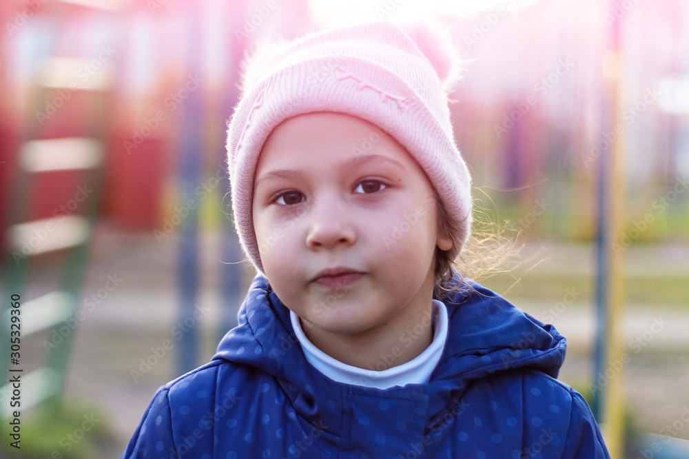 Baby girl in pink hat and blue jacket on playground in rays of setting sun. Portrait. Front view.. Soft focus. Baby girl in pink hat and blue jacket on playground in rays of setting sun. Portrait. Front view.. Soft focus.