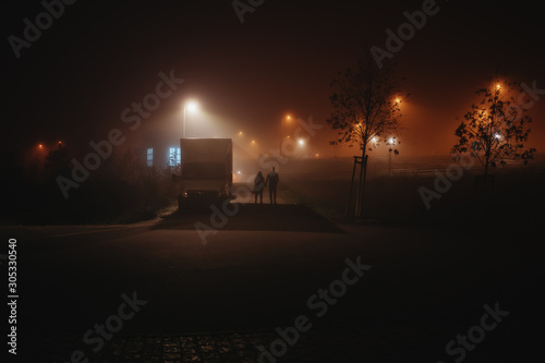 two people walking down a street surrounded by dramatic lighting during a foggy night