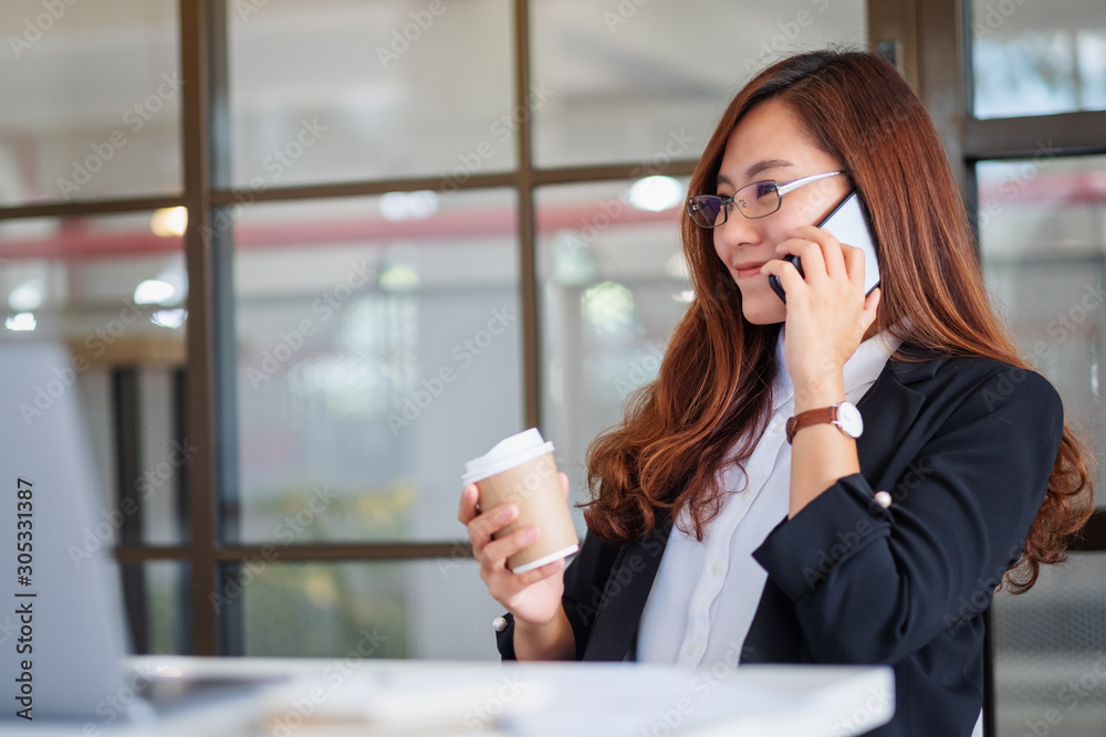Closeup image of a businesswoman holding a cup of coffee while talking on mobile phone in office