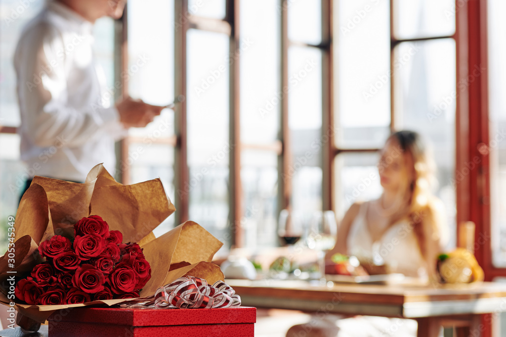Bouquet of fresh red roses and a gift, waiter talking to female customer sitting at restaurant table in background