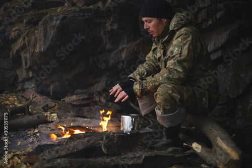 Man in camouflage drinking tea and warms himself by the fire in bad weather. Extreme travel. Survival in the wild.