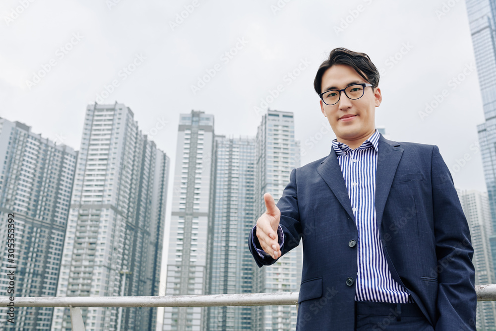 Smiling Asian businessman outstretching hand for handshake, skyscrapers in background Smiling Asian businessman outstretching hand for handshake, skyscrapers in background