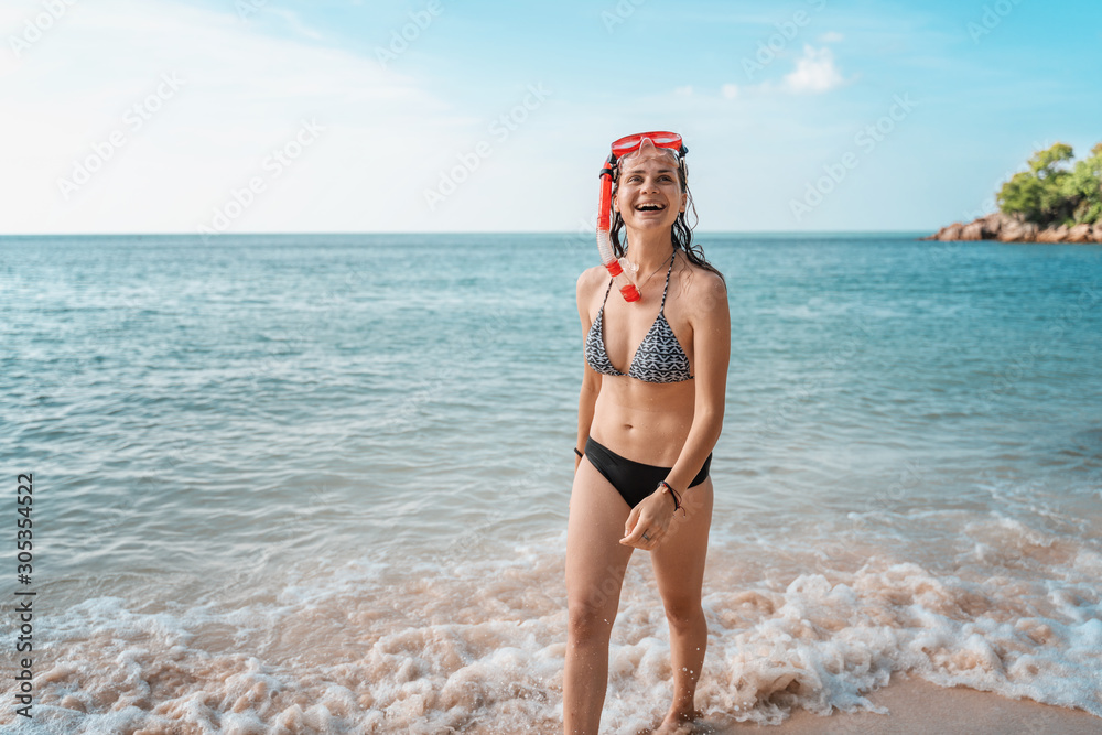 Young happy girl in bikini and with snorkeling mask laughs joyfully and walks along the beach in the tropics, vacation travel sport and entertainment concept Young happy girl in bikini and with snorkeling mask laughs joyfully and walks along the beach in the tropics, vacation travel sport and entertainment concept