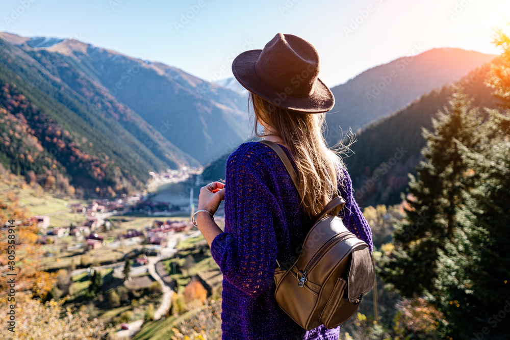 Stylish trendy hipster woman traveler in a felt hat with brown backpack stands on the background of the mountains and uzungol lake in Trabzon during Turkey travel - obrazy, fototapety, plakaty 