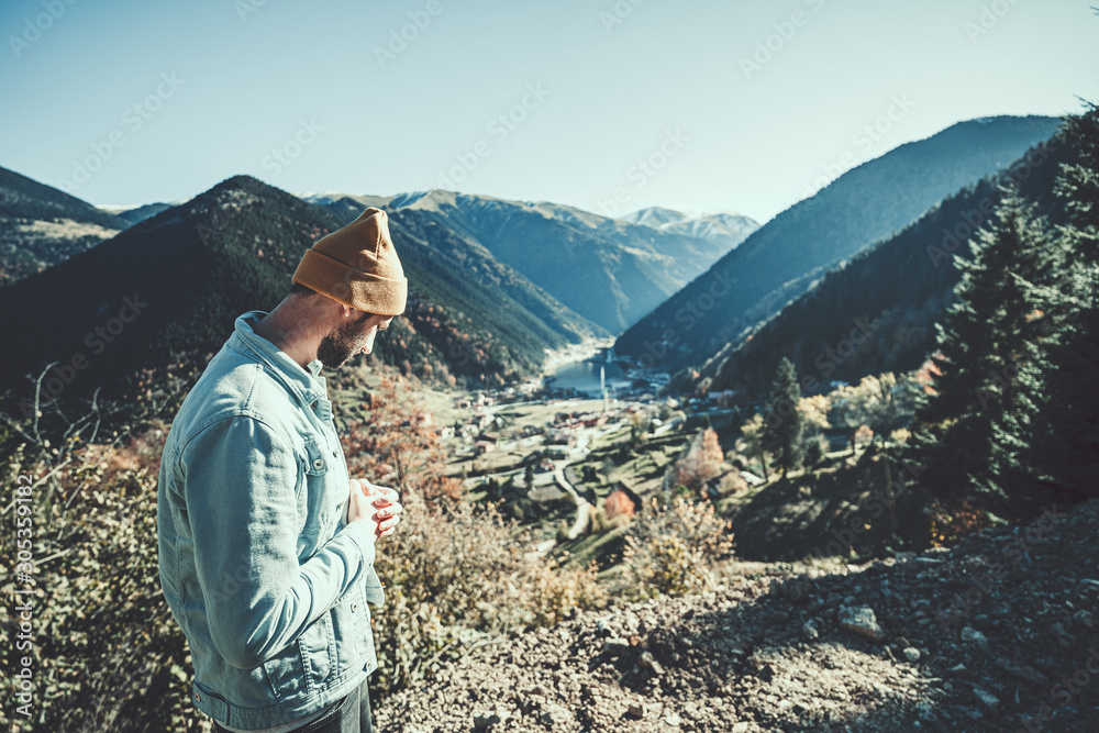 Naklejka premium Stylish trendy hipster man traveler in a yellow hat and denim jacket stands on a background of the mountains and uzungol lake in Trabzon during Turkey travel