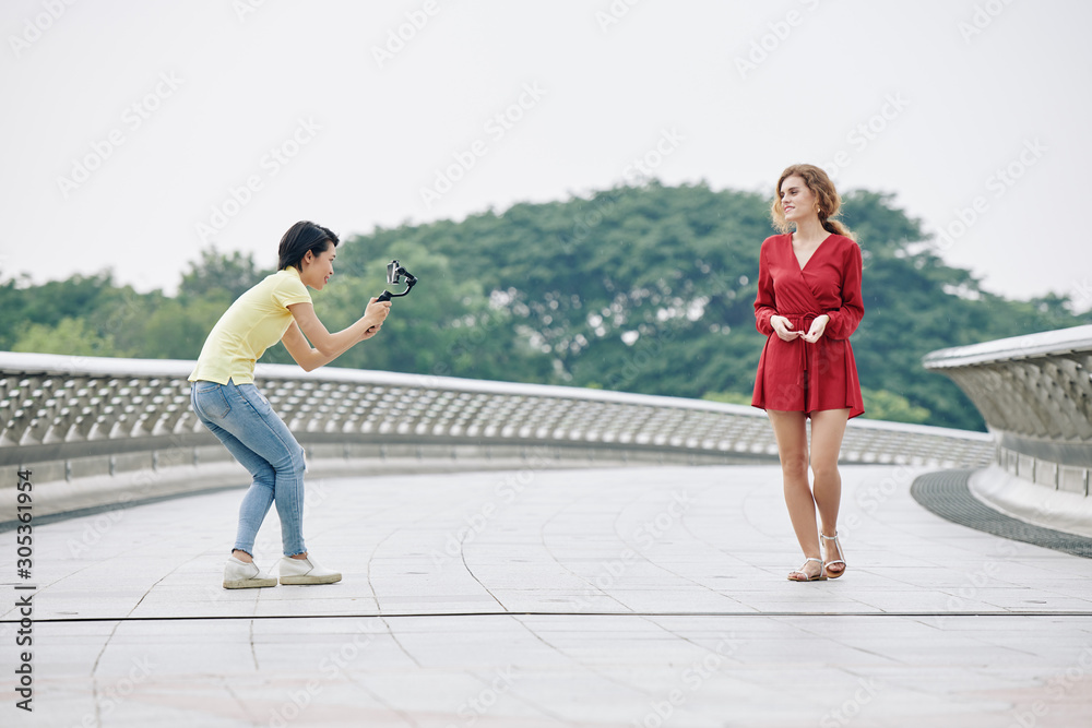 Fototapeta premium Young Vietnamse woman with smartphone filming pretty blogger talking when walking along the bridge