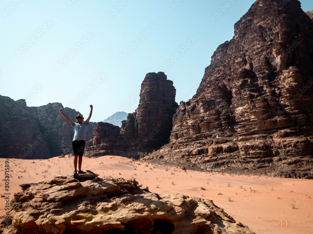 Guy standing on the edge of a cliff in the desert of Wadi Rum with mountains in the background viewed from the top of the cliff