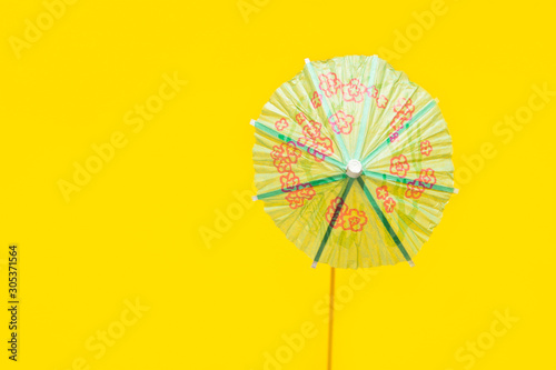 Green paper cocktail umbrella on bright yellow background. Harsh light hard shadow. Trendy minimalist pop art style. Summer vacation travel beach party concept. Poster with copy space