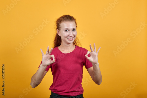 Attractive young lady in red shirt showing okay gesture, smiling isolated on orange background in studio. People sincere emotions, health concept.
