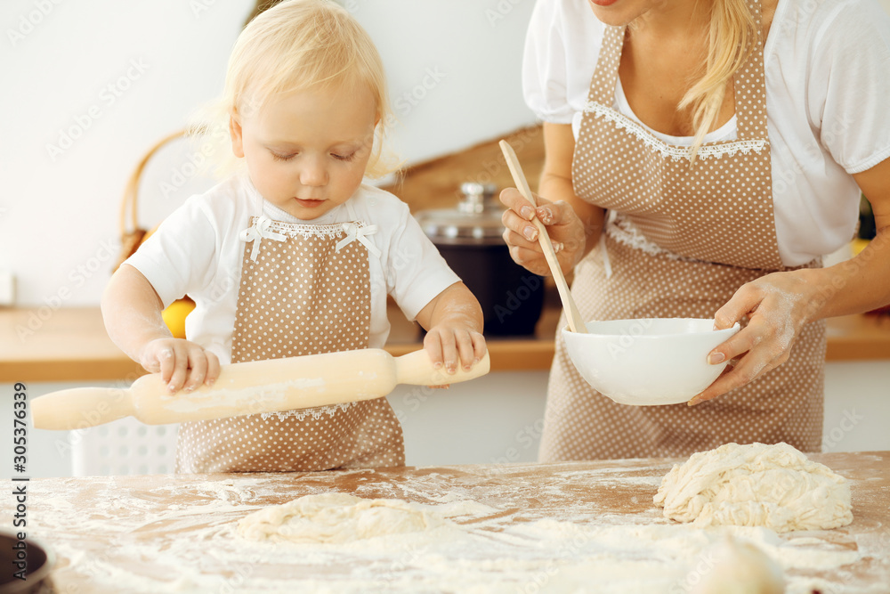 Little girl and her blonde mom in beige aprons playing and laughing while kneading the dough in kitchen. Homemade pastry for bread, pizza or bake cookies. Family fun and cooking concept Little girl and her blonde mom in beige aprons playing and laughing while kneading the dough in kitchen. Homemade pastry for bread, pizza or bake cookies. Family fun and cooking concept