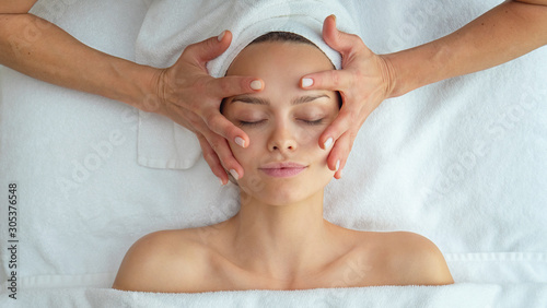 Close up portrait of a beautiful young smiling woman with a towel on her head is receiving a facial massage and spa treatment for perfect skin in a luxury wellness center.