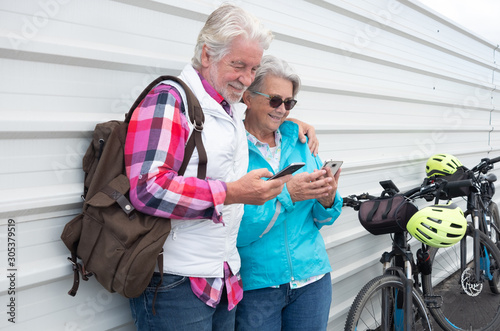 A senior couple with gray hair standing against a white metal wall near to their electric bicycles. Using mobile phone and smiling