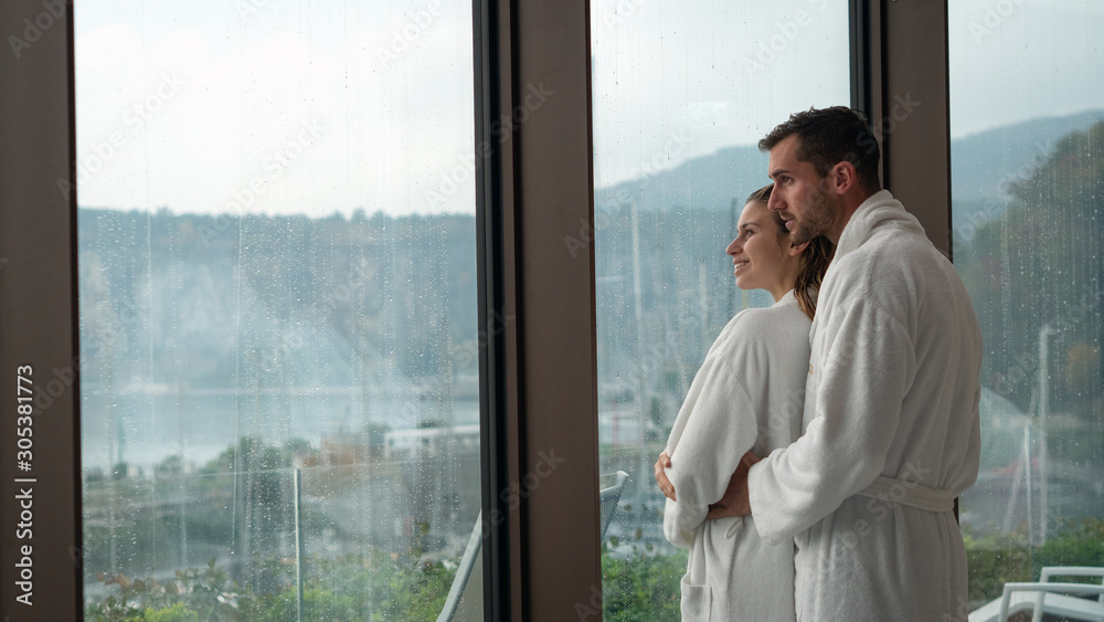 An young happy embraced couple in white bathrobes after various treatments are enjoying the panoramic view through a window in a luxury wellness center An young happy embraced couple in white bathrobes after various treatments are enjoying the panoramic view through a window in a luxury wellness center