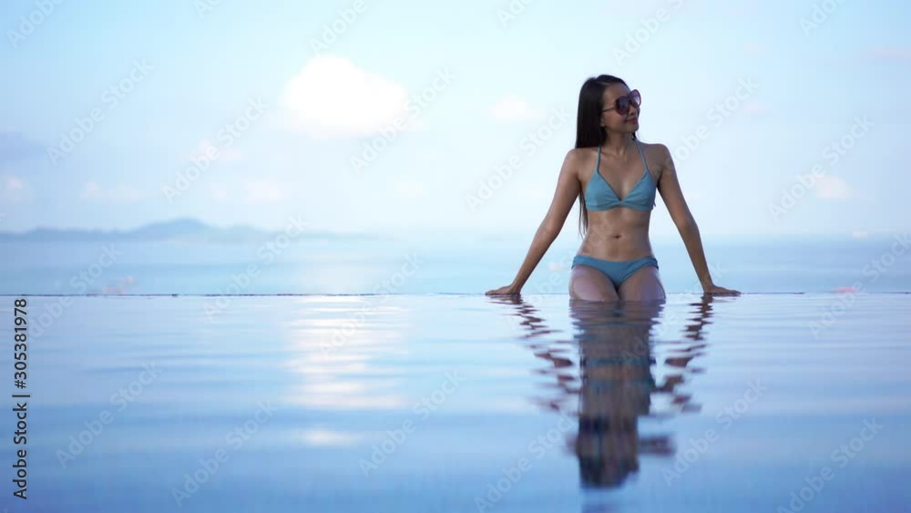 Pretty, fit, young, Asian woman in a blue bikini sits on the edge of a resort infinity pool enjoying the warm tropical sun. Pretty, fit, young, Asian woman in a blue bikini sits on the edge of a resort infinity pool enjoying the warm tropical sun.