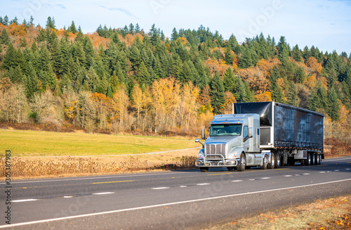 Big rig bonnet semi truck with grille guard transporting cargo in black covered semi trailer moving on the road with autumn trees and meadow