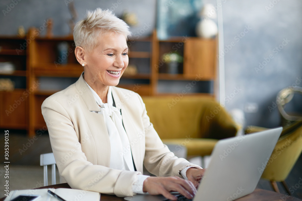 Picture of stylish cheerful middle aged female in elegant suit enjoying online communication while working in office, sitting at desk in front of open portable computer, keyboarding and smiling - obrazy, fototapety, plakaty 