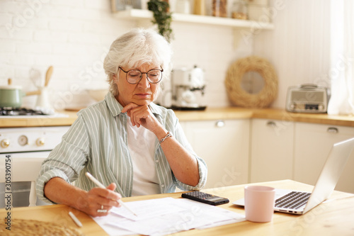 Concentrated female pensioner wearing eyeglasses focused on financial papers while paying bills online using laptop, holding pencil, making notes. People, technology, finances and domestic budget photo