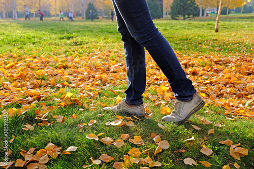 Close up of a woman legs walking in an autumn park on a green lawn among fallen golden leaves; in the background blurred out of focus figures of people