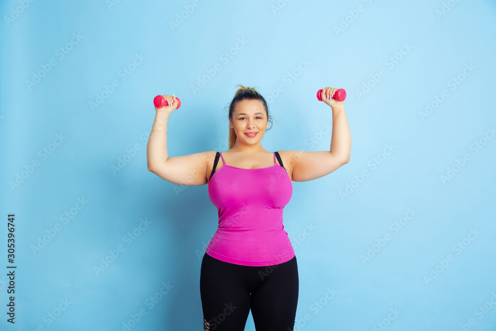 Young caucasian plus size female model's training on blue background. Concept of sport, human emotions, expression, healthy lifestyle, body positive, equality. Training with the weights, copyspace. Young caucasian plus size female model's training on blue background. Concept of sport, human emotions, expression, healthy lifestyle, body positive, equality. Training with the weights, copyspace.