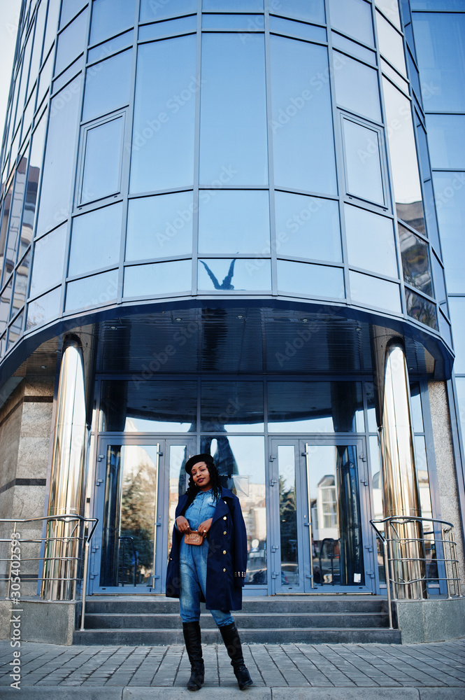 Stylish fashionable african american women in jeans wear and black beret with blue coat against modern building.