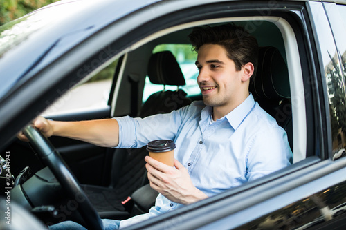 Handsome young man driving a car and holding paper cup of coffee