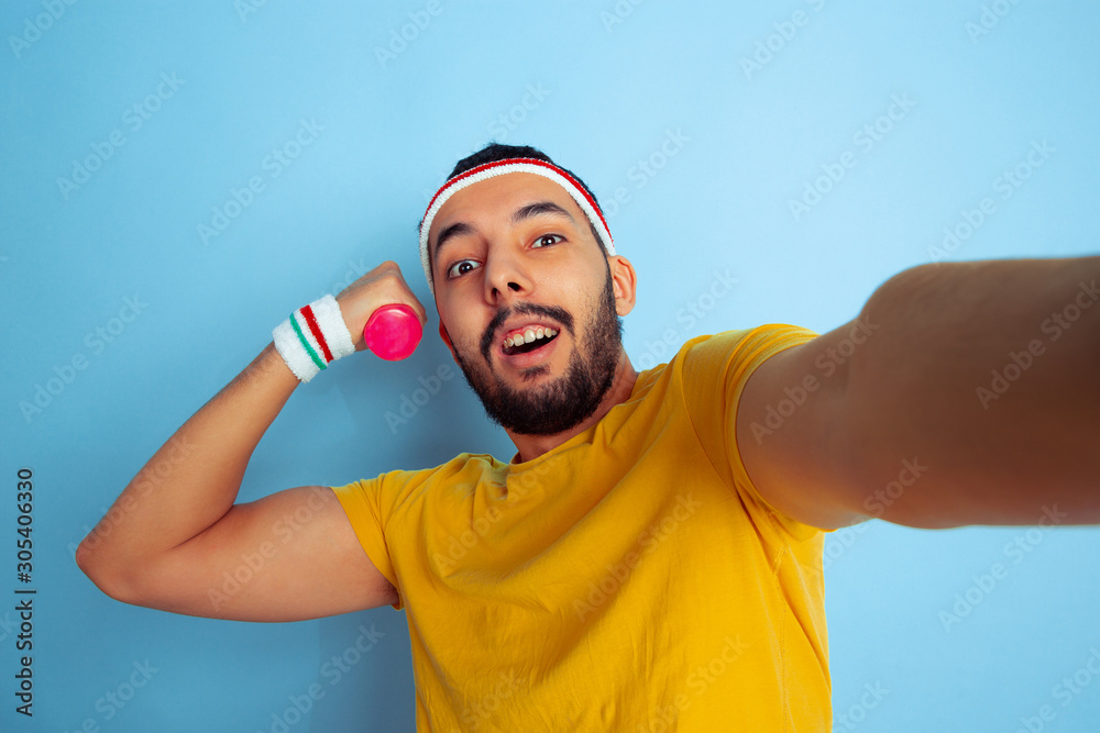 Young caucasian man in bright clothes training on blue background Concept of sport, human emotions, facial expression, healthy lifestyle, youth, sales. Makes selfie while training with the weights.