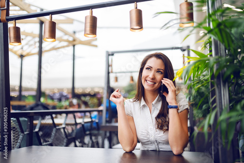 Portrait of gorgeous Caucasian brunette sitting in cafe in shopping mall and gossiping over phone.