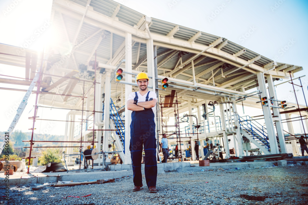 Full length of handsome caucasian worker in overalls and helmet on head standing outdoors with arms crossed. Oil production.