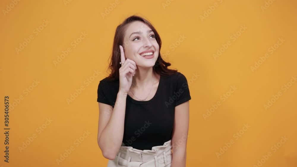 Pretty caucasian young woman in black shirt, keeping hand on waist, pointing directly isolated on orange background in studio. People sincere emotions, health concept. Pretty caucasian young woman in black shirt, keeping hand on waist, pointing directly isolated on orange background in studio. People sincere emotions, health concept.