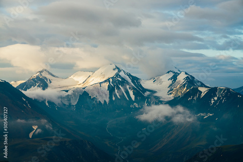 Atmospheric alpine landscape with big snowy mountains among low clouds and highland valley with meltwater stream in golden hour. Wonderful scenery to massive glacier on giant mountain range in sunrise