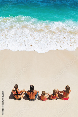 On sunny beach happy kids with parents have fun, sit on white sand see at sea surf with foam and splashes. Top view. Active children lifestyle, summer family vacation travel on tropical family resort.