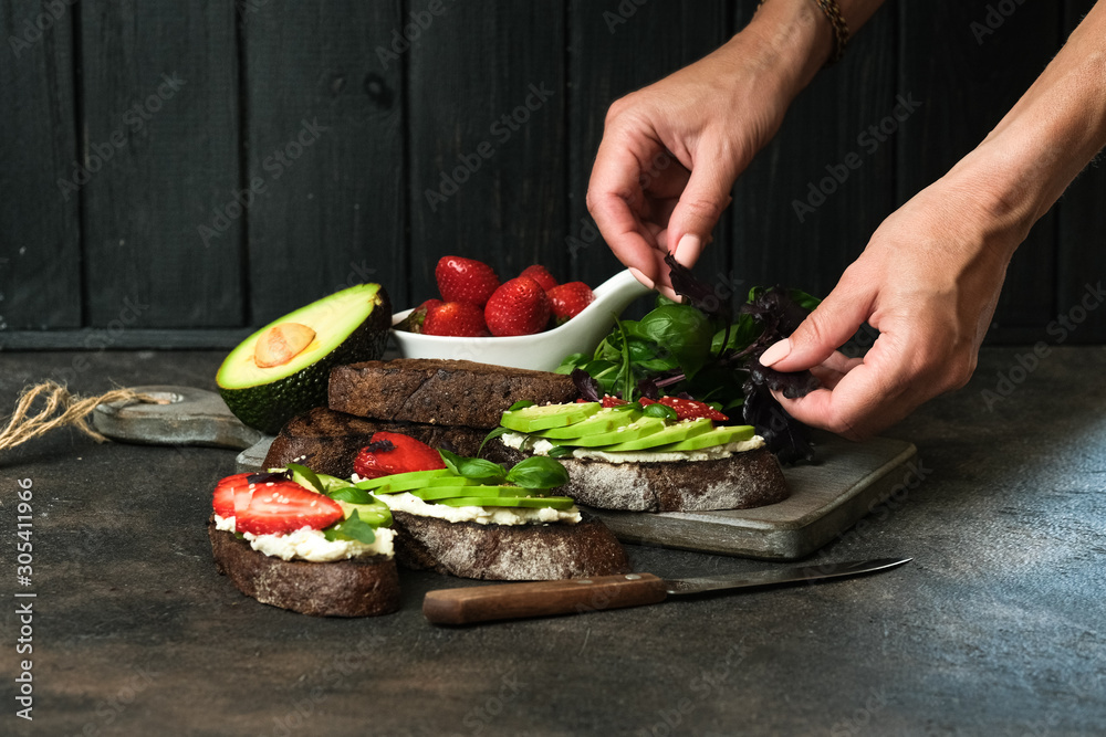 Toast or sandwich with avocado, cheese, strawberries, herbs and seeds on a dark background. Female hands serve a dish. An idea for bruschetta or for a healthy snack. Healthy vegan breakfast.