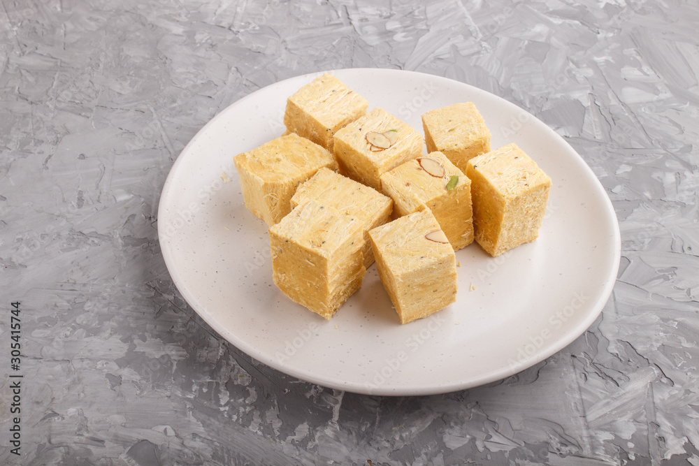 traditional indian candy soan papdi in white plate with almond and pistache on a gray concrete background. side view