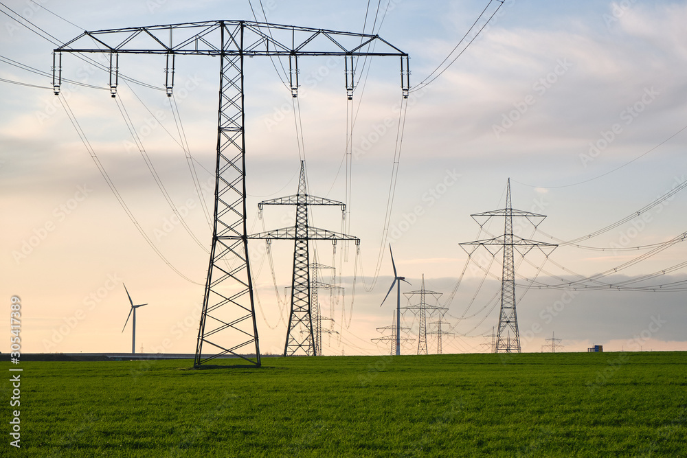 Stromtrasse, Hochspannungsmasten hintereinander aufgereiht bis zum Horizont auf einer grünen Wiese, im Hintergrund Windräder