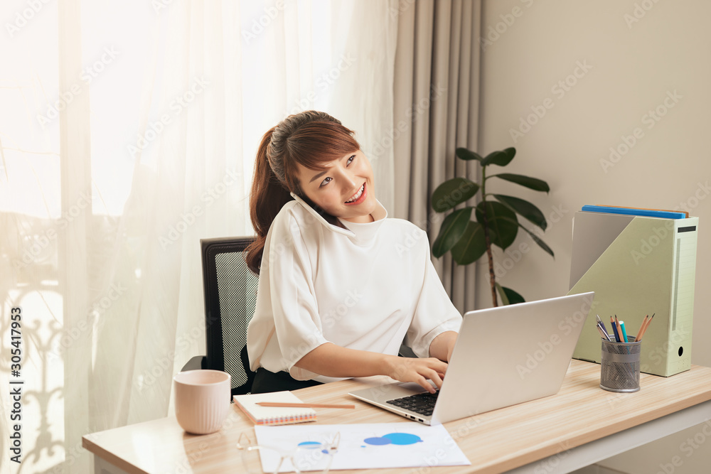 Confident Asian woman talking on her mobile phone and using laptop while sitting at desk and working from home. Home office. Confident Asian woman talking on her mobile phone and using laptop while sitting at desk and working from home. Home office.