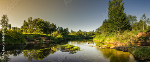 Fototapeta Naklejka Na Ścianę i Meble -  Tranquil summer evening landscape. panoramic view of the river with lush coastal vegetation, snags in the water, and sunset glow on the left