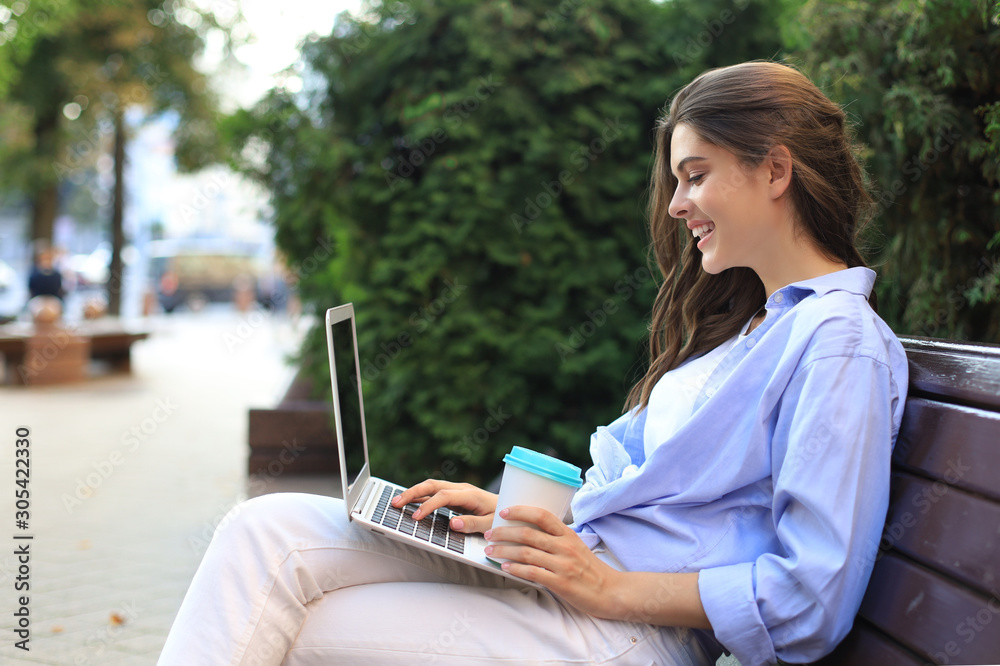 Beautiful woman sitting on bench holding coffee while using laptop outdoor. Beautiful woman sitting on bench holding coffee while using laptop outdoor.