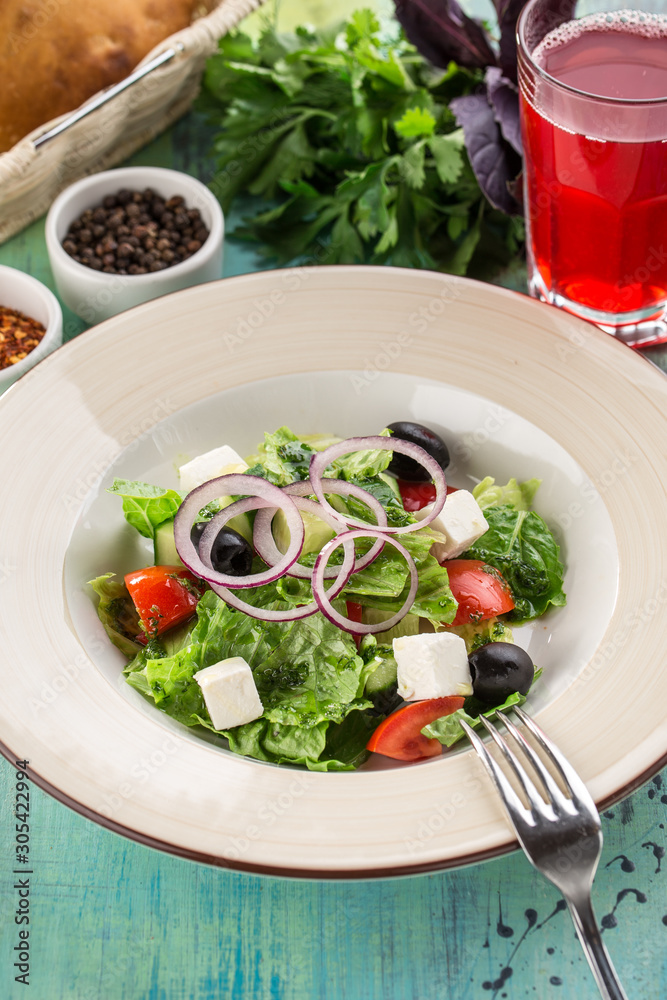 Fresh Greek salad with olives, tomato, feta cheese, cucumber and onion and glass of red drink on blue wooden table Fresh Greek salad with olives, tomato, feta cheese, cucumber and onion and glass of red drink on blue wooden table