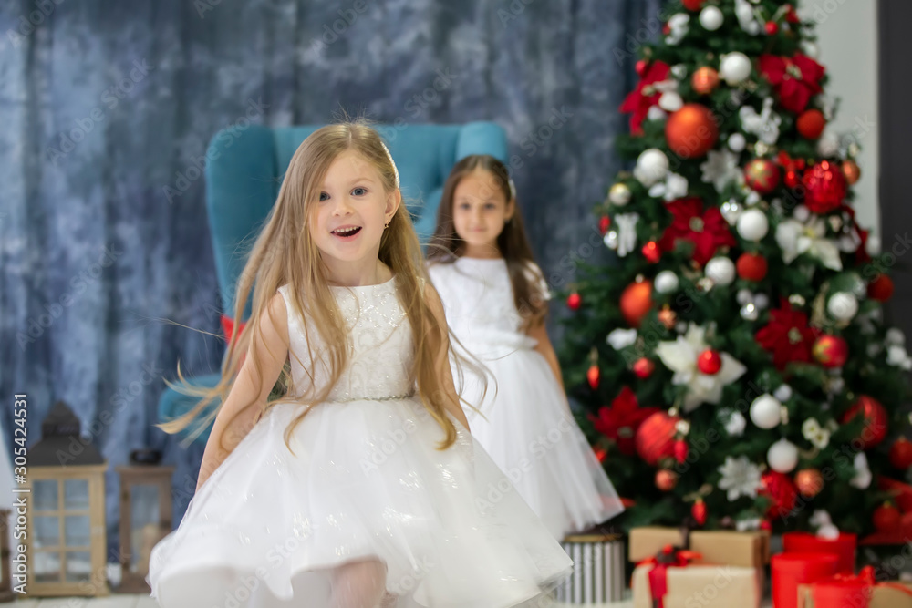 Merry children at the Christmas holiday. Two little girls in elegant dresses against the background of the Christmas tree Merry children at the Christmas holiday. Two little girls in elegant dresses against the background of the Christmas tree