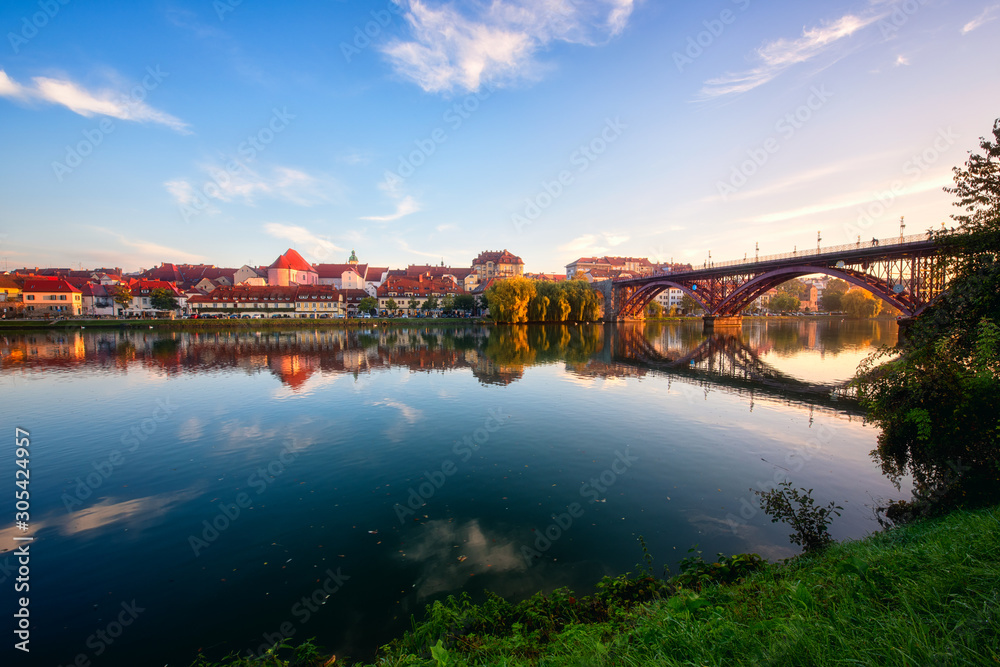 Fototapeta premium Amazing view of Maribor Old city, Main bridge (Stari most) on the Drava river at sunrise, Slovenia. Scenic cityscape with blue sky and reflection, travel background for wallpaper or guide book