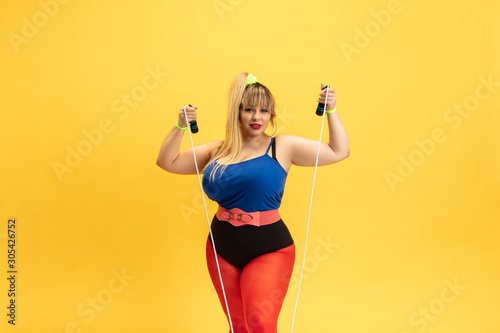 Young caucasian plus size female model's training on yellow background. Stylish woman in bright clothes. Copyspace. Concept of sport, healthy lifestyle, body positive, fashion. Posing with jump rope.