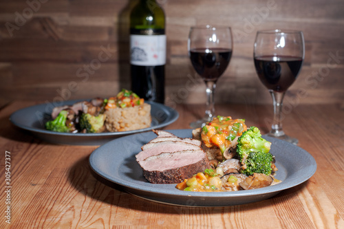 Two plates with duck breast and vegetables on wooden table with glasses of red wine