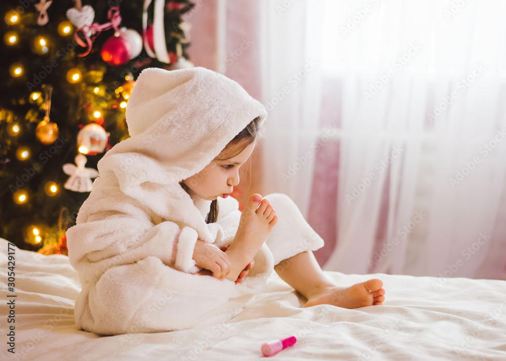 Fototapeta premium A little girl paints her nails. Girl sitting near the Christmas tree on the bed and paints her nails. Preparing to new year party
