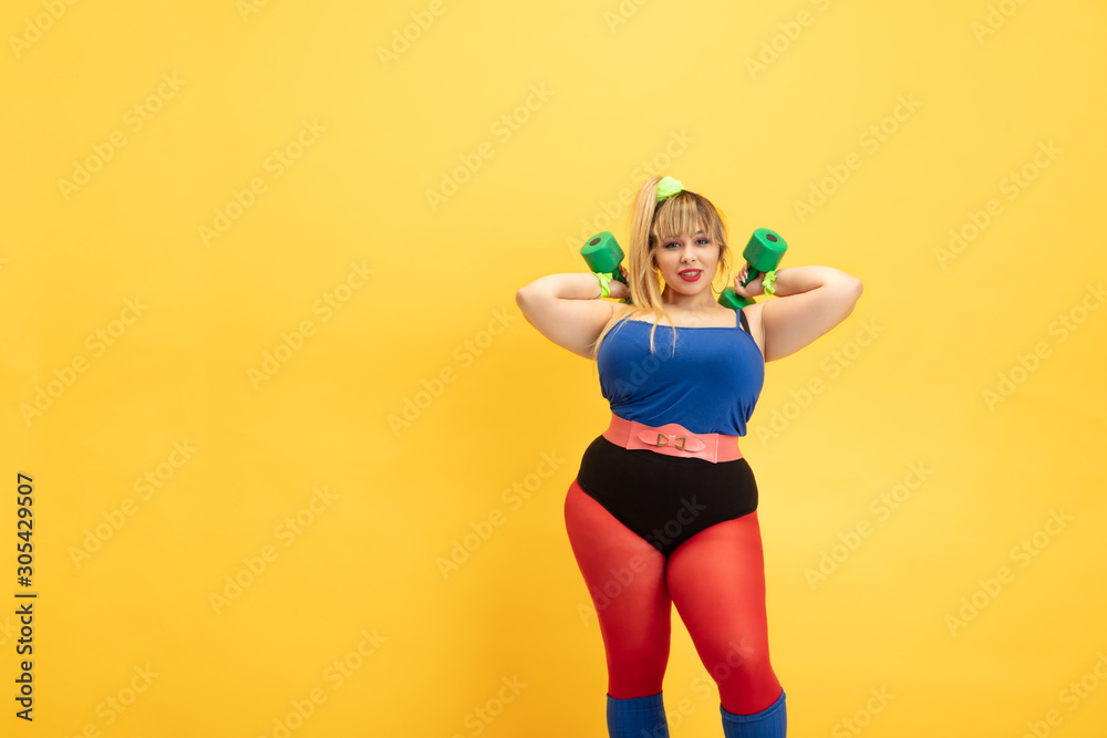 Young caucasian plus size female model's training on yellow background. Stylish woman in bright clothes. Copyspace. Concept of sport, healthy lifestyle, body positive, fashion. Practicing with weights Young caucasian plus size female model's training on yellow background. Stylish woman in bright clothes. Copyspace. Concept of sport, healthy lifestyle, body positive, fashion. Practicing with weights