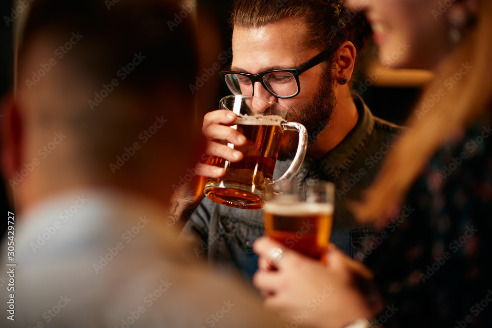 Handsome bearded caucasian young man with eyeglasses standing in pub with friends and drinking fresh beer. Handsome bearded caucasian young man with eyeglasses standing in pub with friends and drinking fresh beer.