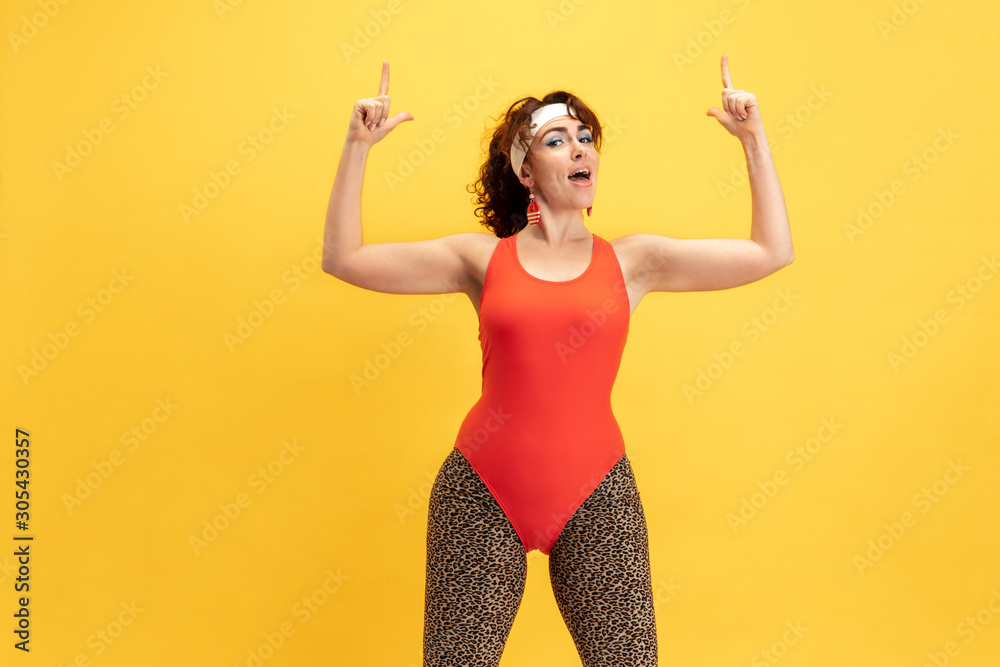 Young caucasian plus size female model's training on yellow background. Stylish woman in bright clothes. Copyspace. Concept of sport, healthy lifestyle, body positive, fashion. Smiling, pointing up. Young caucasian plus size female model's training on yellow background. Stylish woman in bright clothes. Copyspace. Concept of sport, healthy lifestyle, body positive, fashion. Smiling, pointing up.