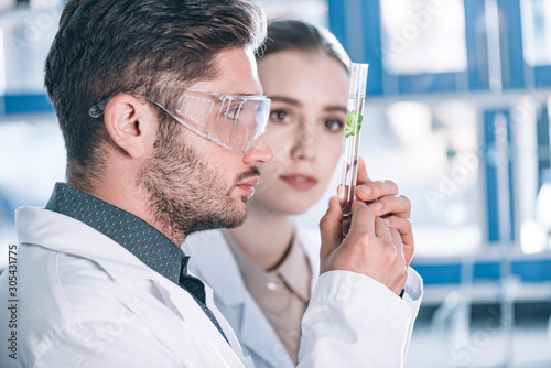 selective focus of handsome scientist in goggles looking at test tube with plant near beautiful coworker