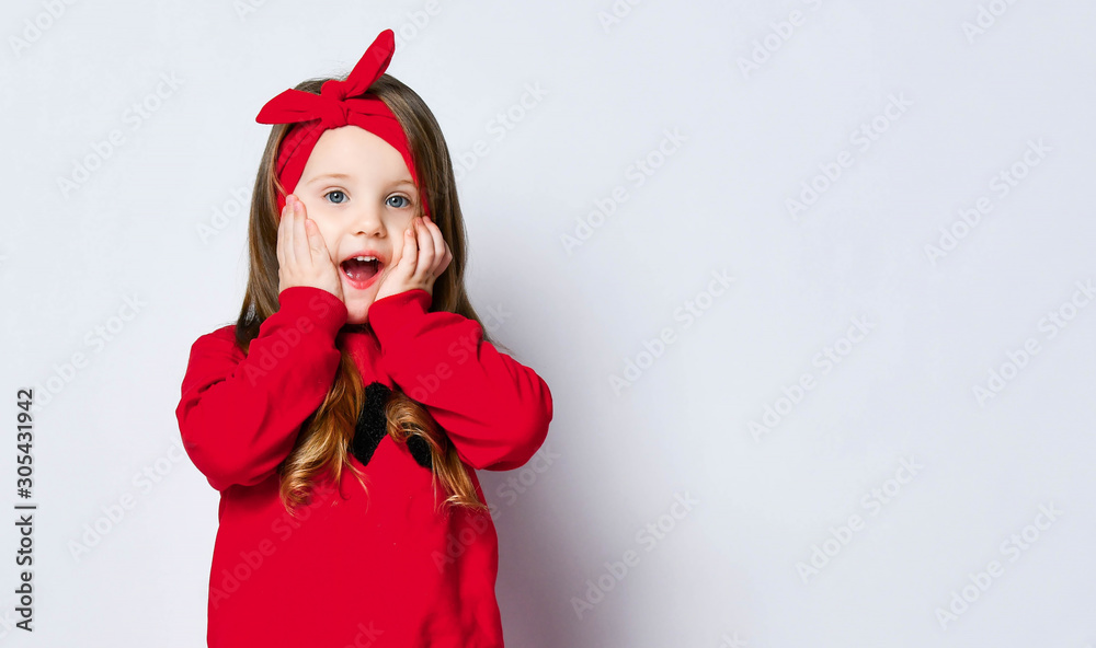 Happy surprised amazed cute little kid girl in red sweater and headband with bow is covering cheeks with her arms Happy surprised amazed cute little kid girl in red sweater and headband with bow is covering cheeks with her arms